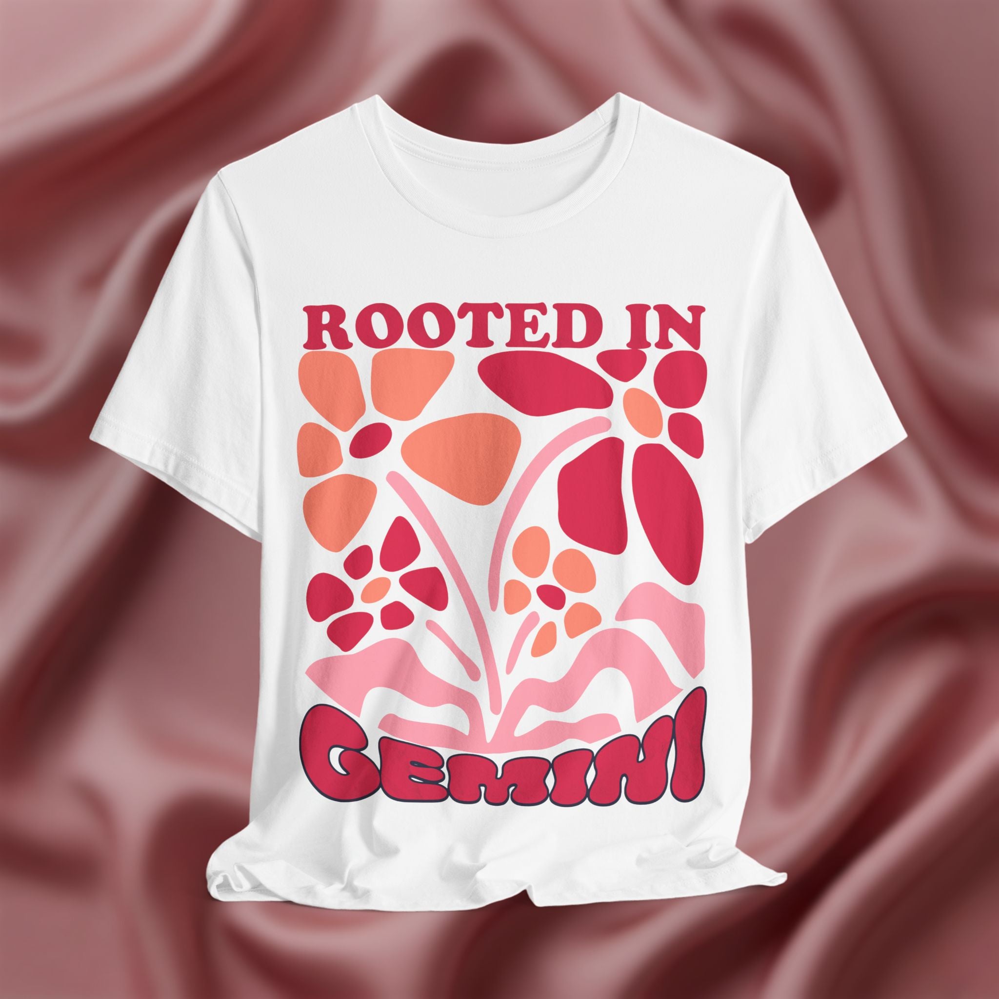 White t-shirt with floral design and text 'Rooted in Gemini' on a maroon fabric background