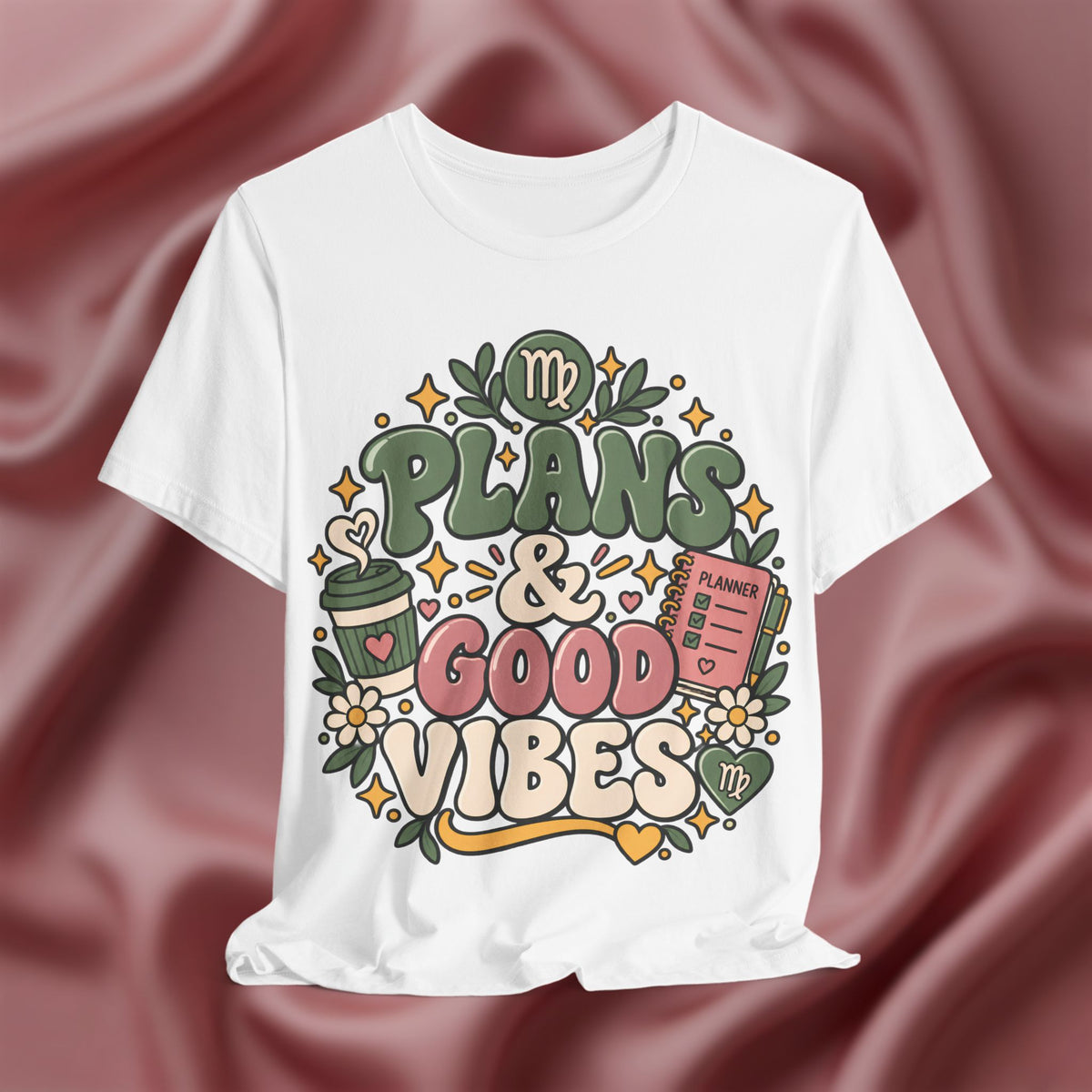 A white short-sleeve graphic T-shirt displayed against a draped dusty rose satin fabric background. The shirt features a colorful retro-style graphic with the text 'PLANS & GOOD VIBES' and various illustrations including a coffee cup, planner, flowers, and stars.