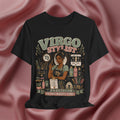 A black graphic t-shirt featuring a detailed illustration of a hair stylist surrounded by salon tools and products, with the text 'VIRGO STYLIST' and 'PRECISION IS THE WHOLE VIBE'. The t-shirt is displayed against a draped, satin-like dusty rose background.