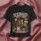 A black graphic t-shirt featuring a detailed illustration of a hair stylist surrounded by salon tools and products, with the text 'VIRGO STYLIST' and 'PRECISION IS THE WHOLE VIBE'. The t-shirt is displayed against a draped, satin-like dusty rose background.