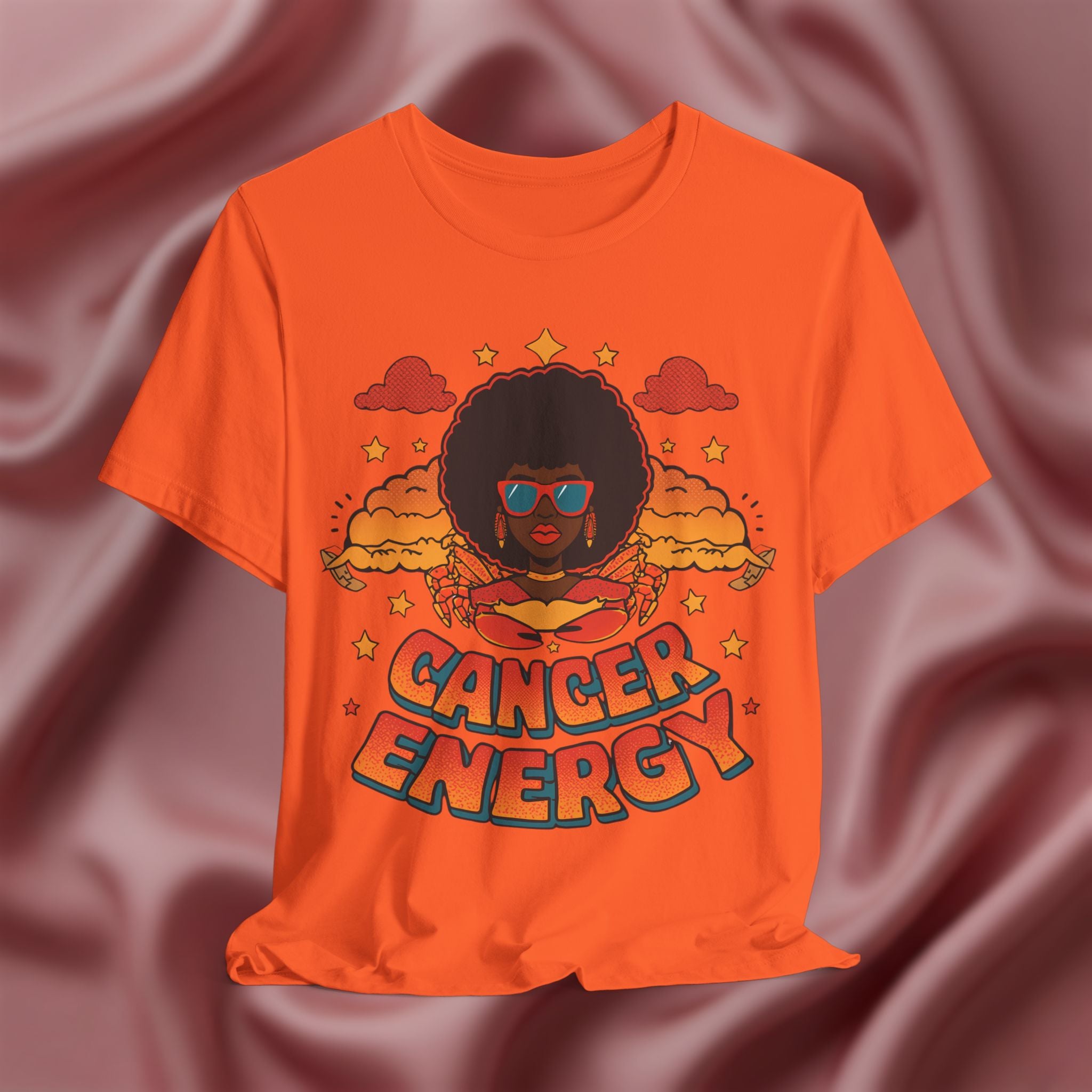 Orange t-shirt with a graphic design of a person with an afro and 'Cancer Energy' text on a maroon fabric background.