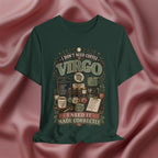 An olive green graphic t-shirt featuring a vintage-style Virgo-themed coffee design with illustrations of coffee beans, mugs, and text reading 'I DON'T NEED COFFEE VIRGO' and 'I NEED IT MADE CORRECTLY', displayed against a draped pink satin background.