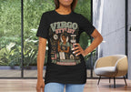 A woman modeling a black graphic t-shirt with a 'Virgo Stylist' design, standing in a modern room with large windows showing green foliage outside.