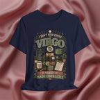 A navy blue graphic t-shirt featuring a vintage-style astrological design for Virgo with coffee-themed illustrations and the text 'I DON'T NEED COFFEE VIRGO I NEED IT MADE CORRECTLY', displayed against a draped pink satin fabric background.
