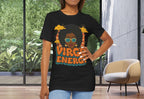 A woman modeling a black graphic t-shirt with a colorful retro design featuring the text 'VIRGINE ENERGY' and an illustration of a woman with sunglasses, standing in a modern room with large windows.
