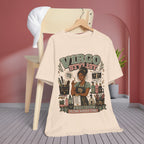 A cream-colored graphic t-shirt hanging on a wooden hanger from a light wood chair, set against a styled room background with a pink wall, gray wood floor, and a white cap draped over the chair.