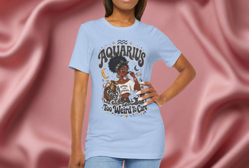 Woman wearing a light blue t-shirt with 'Aquarius Too Weird To Care' design featuring a black woman wearing a t-shirt with the text 'different is my superpower' on a pink fabric background