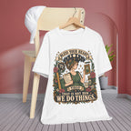 A white graphic t-shirt hanging on a wooden hanger in front of a light wood chair, set against a styled background with a pinkish-red wall, gray wood floor, and a white cap draped over the chair.