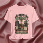 A light pink short-sleeve t-shirt with a vintage-style graphic print featuring the text 'I DON'T NEED COFFEE VIRGO' and 'I NEED IT MADE CORRECTLY' with illustrations of coffee cups, mugs, and brewing equipment. The t-shirt is displayed against a draped, satin-like dusty rose background.