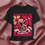 Black t-shirt with colorful floral design and 'Rooted in Gemini' text on a pink fabric background
