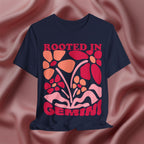 Navy blue t-shirt with colorful floral design and 'Rooted in Gemini' text on a pink fabric background