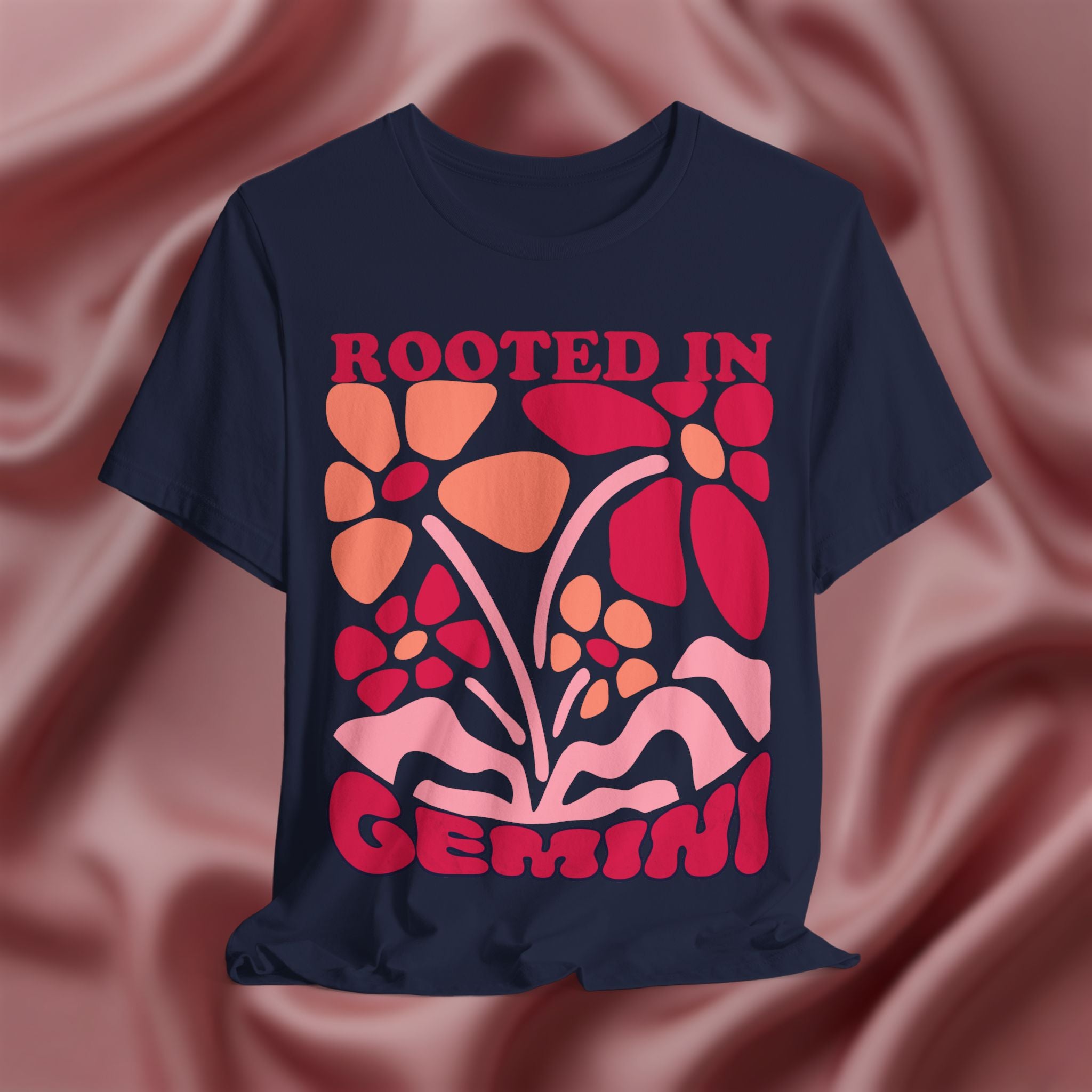 Navy blue t-shirt with colorful floral design and 'Rooted in Gemini' text on a pink fabric background