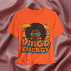 An orange short-sleeve t-shirt with a retro-style graphic of a woman with an afro, sunglasses, and the text 'VIRGO ENERGY' in colorful bubble letters, displayed against a draped mauve satin fabric background.