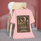 A light pink short-sleeve T-shirt hanging on a wooden hanger against a pink wall, with a stylized vintage-style 'The Maiden Virgo' graphic printed on the front. A wooden chair with a white shirt draped over it and a potted plant are visible in the background.
