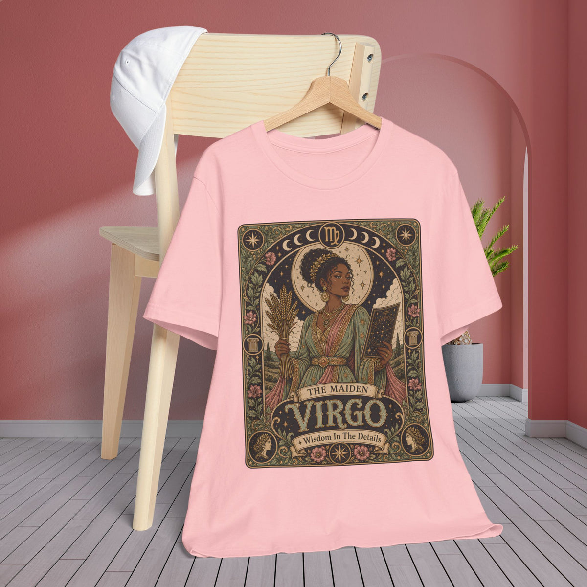 A light pink short-sleeve T-shirt hanging on a wooden hanger against a pink wall, with a stylized vintage-style 'The Maiden Virgo' graphic printed on the front. A wooden chair with a white shirt draped over it and a potted plant are visible in the background.