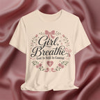 Soft cream short-sleeve t-shirt with a graphic print reading 'Girl, Breathe God Is Still In Control' in black script, surrounded by pink bows, green leaves, and small flowers, displayed against a draped dusty rose satin background.