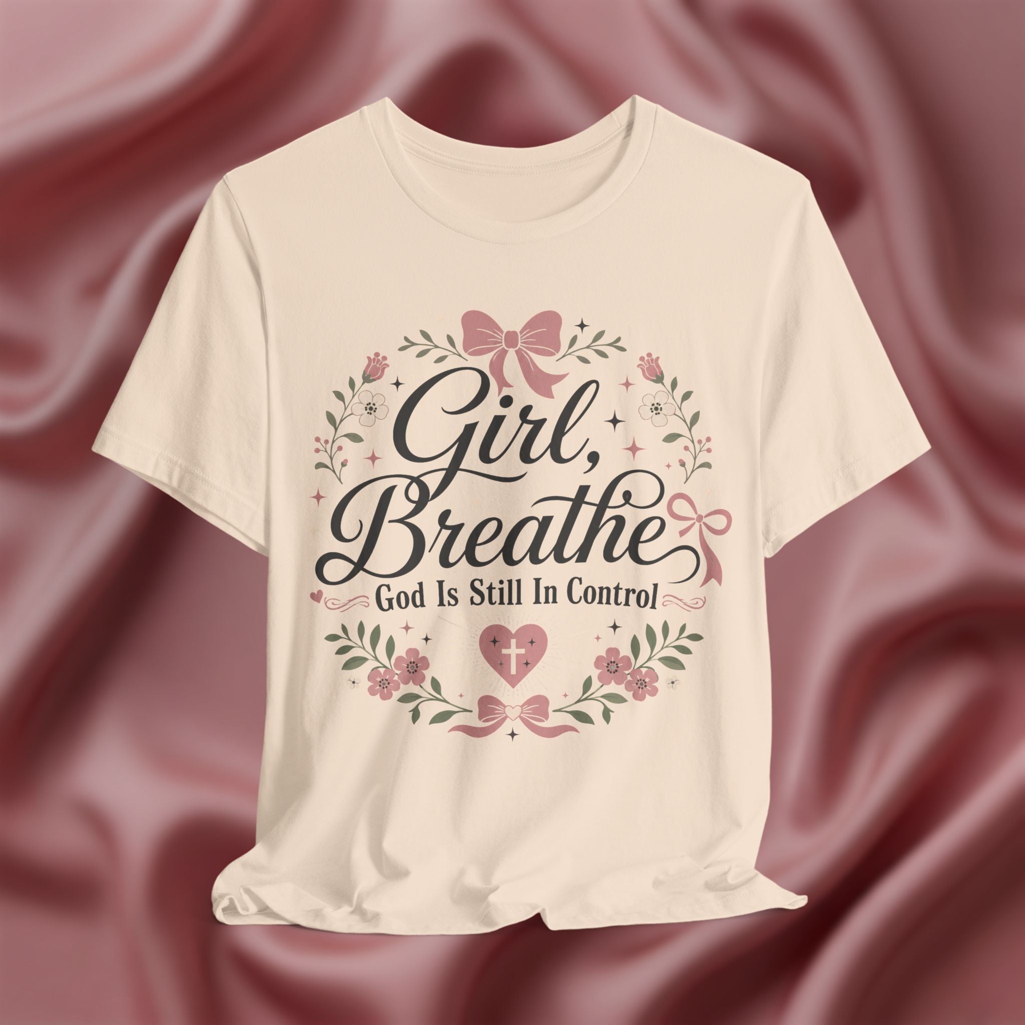 Soft cream short-sleeve t-shirt with a graphic print reading 'Girl, Breathe God Is Still In Control' in black script, surrounded by pink bows, green leaves, and small flowers, displayed against a draped dusty rose satin background.