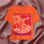 Orange t-shirt with floral design and text 'Rooted in Genius' on a maroon fabric background