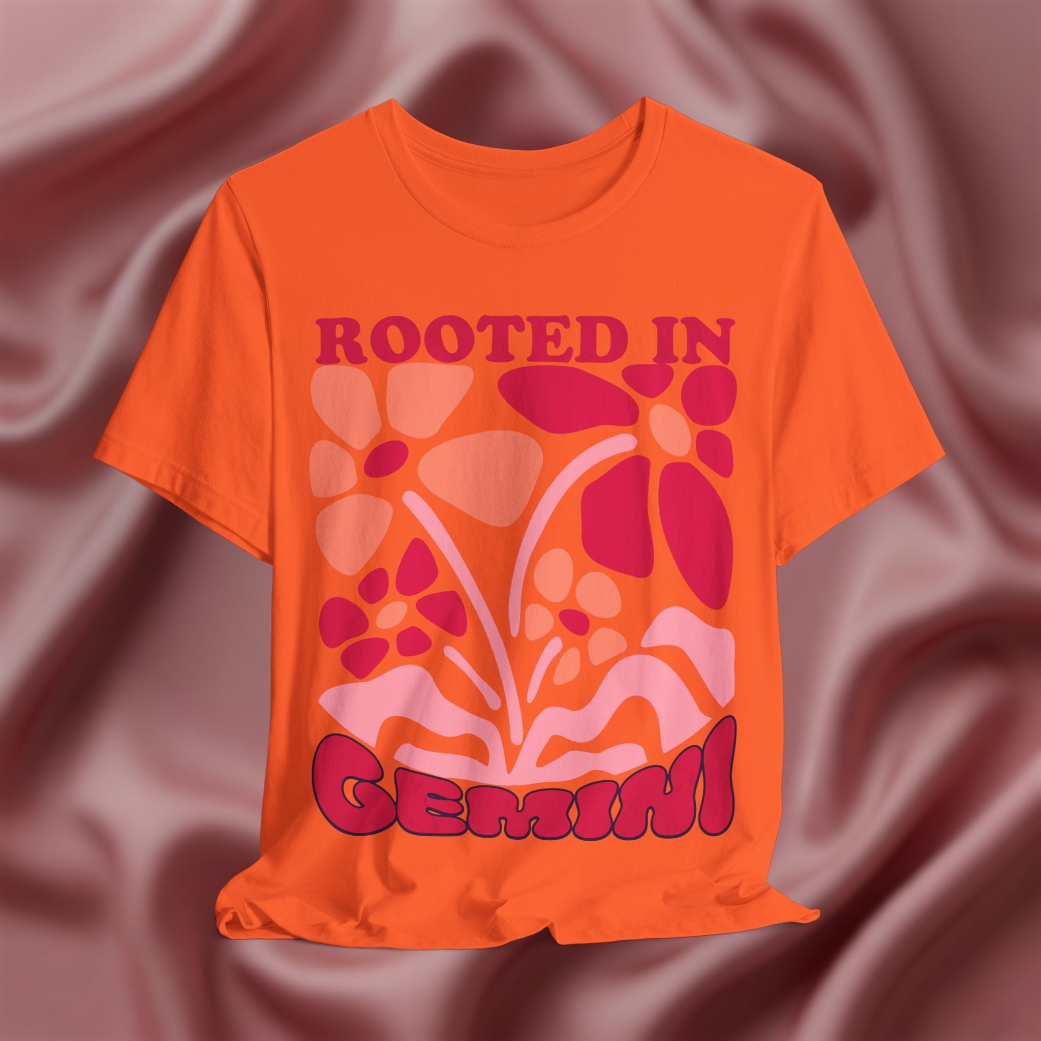 Orange t-shirt with floral design and text 'Rooted in Genius' on a maroon fabric background