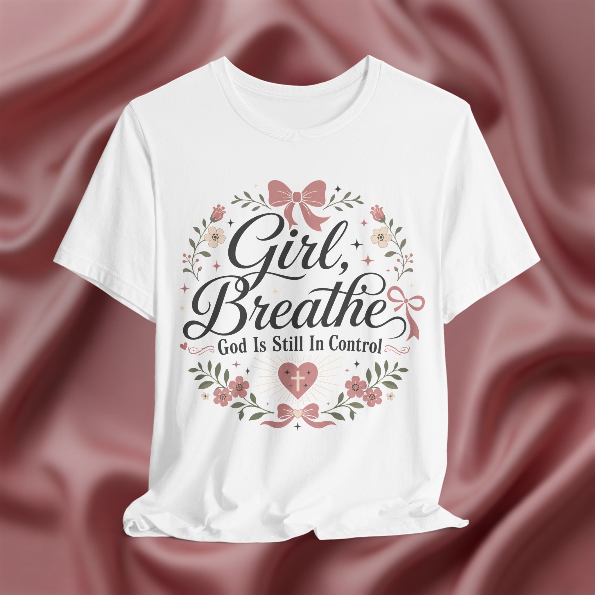 A white short-sleeve crewneck t-shirt with a graphic print reading 'Girl, Breathe God Is Still In Control' in black script, decorated with pink bows, green leaves, and small flowers, displayed against a draped dusty rose satin background.