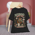 A black graphic t-shirt hanging on a wooden hanger from a light wood chair, set against a pink wall and gray wood floor. The graphic features a detailed illustration of a Virgo woman surrounded by tools, with the text 'Virgo Stylist' and 'Precision is the whole vibe'.