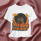 A white short-sleeve t-shirt with a retro-style graphic of a Black woman with an afro, sunglasses, and the text 'VIRGO ENERGY' in orange and teal, displayed against a draped dusty rose satin fabric background.