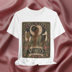 A white short-sleeve crewneck t-shirt featuring a detailed vintage-style graphic of 'The Maiden Virgo' on the front, displayed against a draped dusty rose satin fabric background.