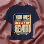 Navy blue t-shirt with 'I'm not a mess, I'm a Gemini' design on a pink fabric background