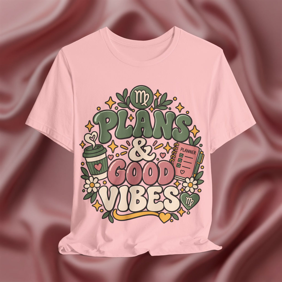 A light pink short-sleeve t-shirt with a colorful graphic print that reads 'PLANS & GOOD VIBES' in green, pink, and cream lettering, surrounded by doodles of a coffee cup, flowers, and a planner, displayed against a draped mauve satin background.