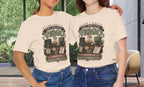 Two women standing indoors wearing matching cream graphic T-shirts with a vintage-style Virgo design and blue jeans, posed in front of large windows with greenery outside.