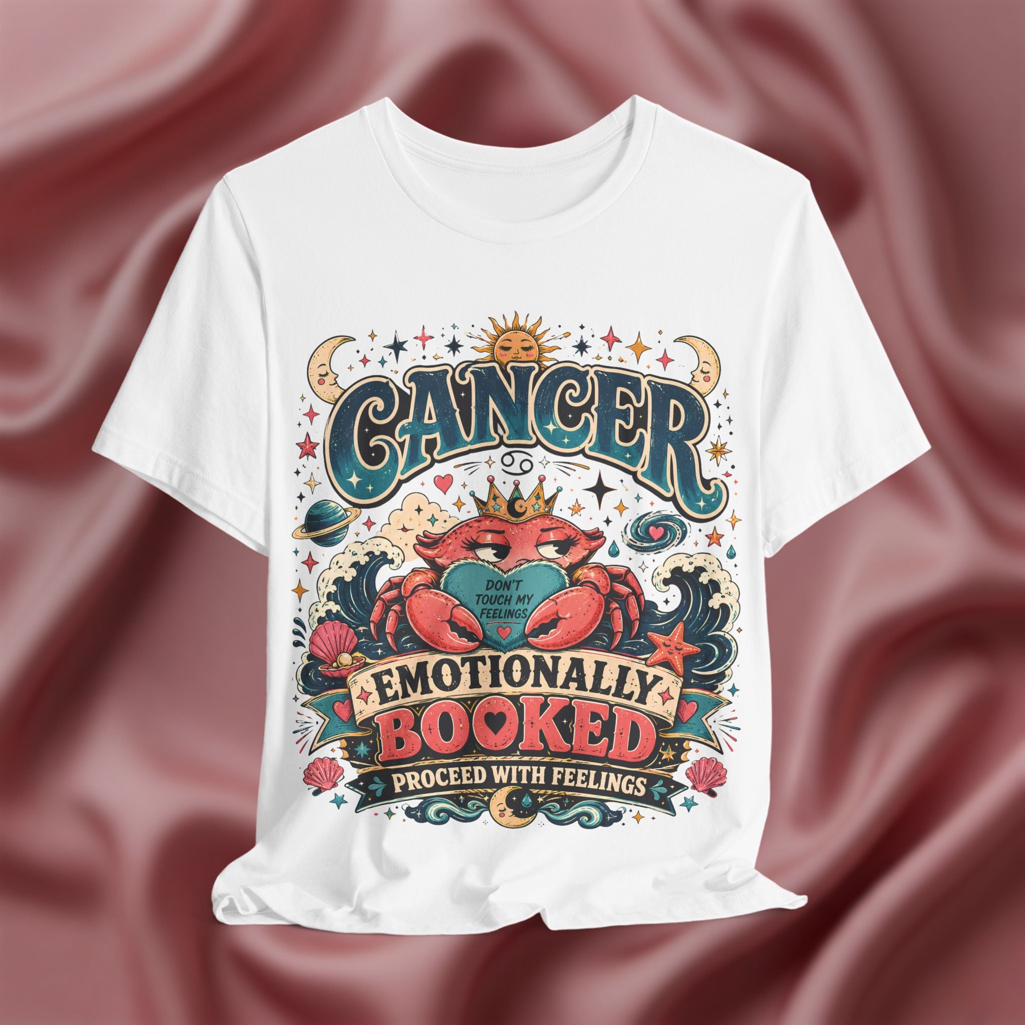 White t-shirt with colorful 'Cancer Emotionally Booked' design on a maroon fabric background