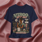 A navy blue graphic t-shirt featuring a detailed illustration of a Virgo stylist surrounded by hair tools and products, with the text 'VIRGO STYLIST' and 'PRECISION IS THE WHOLE VIBE'. The t-shirt is displayed against a draped, satin-like dusty rose background.