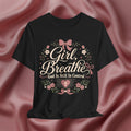 A black short-sleeve graphic T-shirt displayed against a draped pink satin fabric background. The shirt features a central graphic with the words 'Girl, Breathe' and 'God Is Still In Control' in white script, surrounded by pink bows, green leaves, and small white flowers.