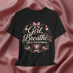 A black short-sleeve graphic T-shirt displayed against a draped pink satin fabric background. The shirt features a central graphic with the words 'Girl, Breathe' and 'God Is Still In Control' in white script, surrounded by pink bows, green leaves, and small white flowers.