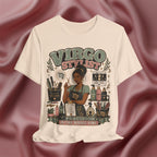 A soft cream graphic t-shirt featuring a vintage-style illustration of a woman styled as a 'Virgo Stylist' with makeup tools and bottles, set against a draped burgundy satin background.