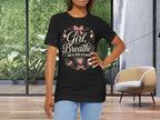 A woman modeling a black graphic t-shirt with the text 'Girl, Breathe God Is Still In Control' surrounded by pink flowers and a bow, set in a modern room with large windows.