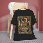 A black short-sleeve graphic T-shirt hanging on a wooden hanger against a pink wall, with a light wood chair and a white cap in the background. The shirt features a large vintage-style astrological print reading 'The Maiden Virgo' with moon phases, a figure in robes, and ornate decorative borders.