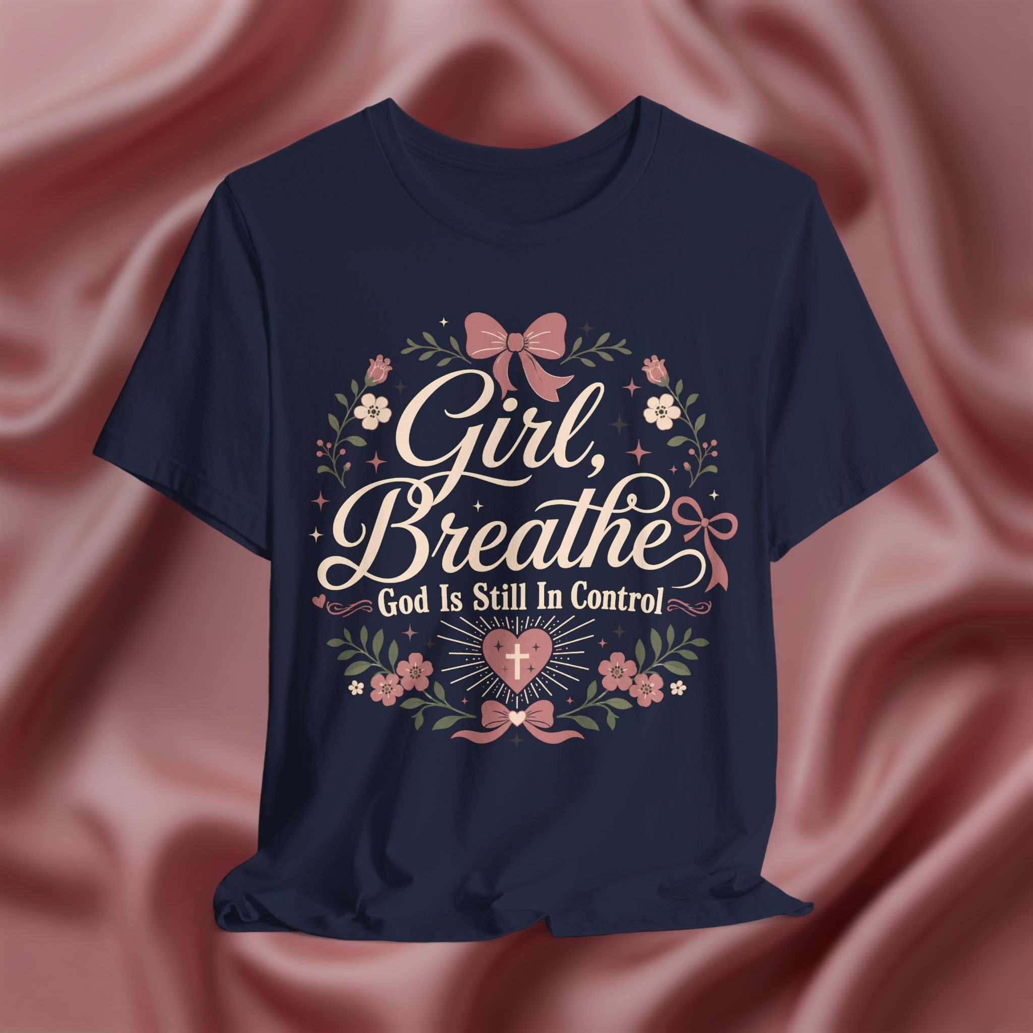 A navy blue short-sleeve t-shirt with a floral graphic and the text 'Girl, Breathe God Is Still In Control' in white script, displayed against a draped pink satin background.