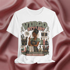 A white graphic t-shirt featuring a detailed illustration of a hairstylist with the text 'VIRGO STYLIST' and 'PRECISION IS THE WHOLE VIBE'. The t-shirt is displayed against a draped, satin-like burgundy background.