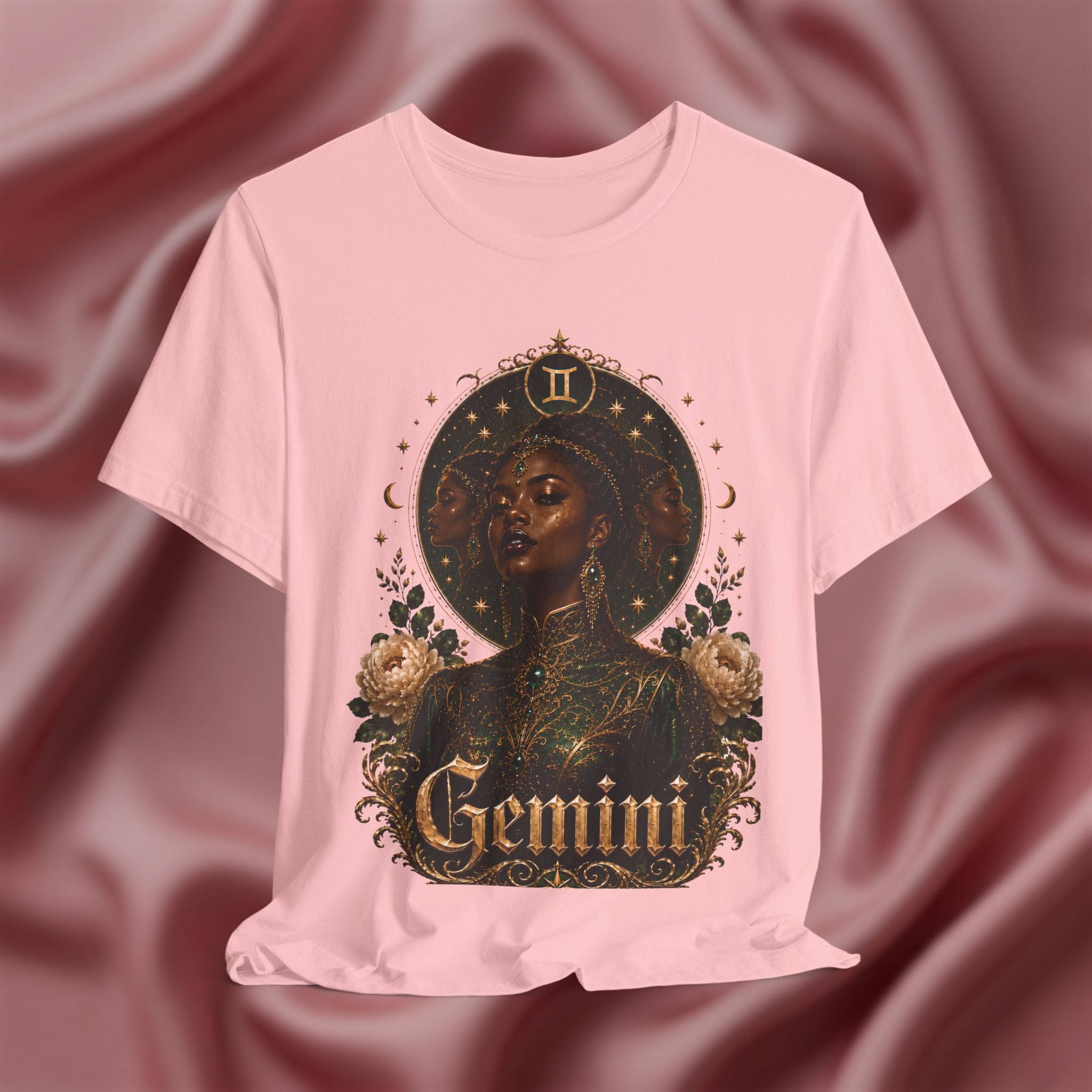 Pink t-shirt with a graphic design of a black woman, flowers, and the word 'Gemini' on a maroon fabric background.