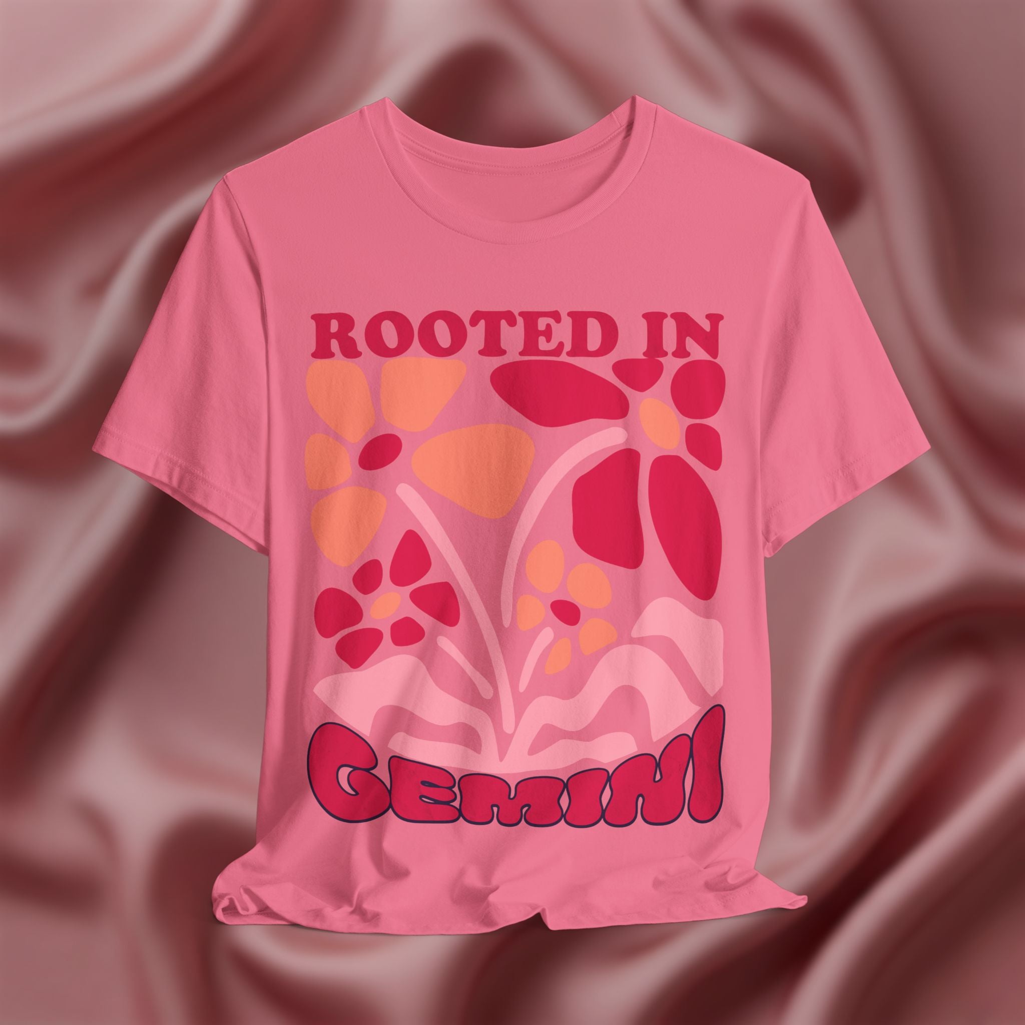 Pink t-shirt with floral design and text 'Rooted in Gemini' on a textured brown background