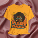 A gold yellow graphic t-shirt featuring a retro-style illustration of a woman with an afro, sunglasses, and the text 'VIRGO ENERGY' in orange and blue, displayed against a draped mauve satin fabric background.