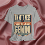 Gray t-shirt with 'I'm not a mess, I'm a Gemini' design on a pink fabric background