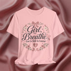 A light pink short-sleeve t-shirt with a graphic print reading 'Girl, Breathe God Is Still In Control' in black script, surrounded by a floral wreath with pink bows and green leaves, displayed against a draped satin fabric background.