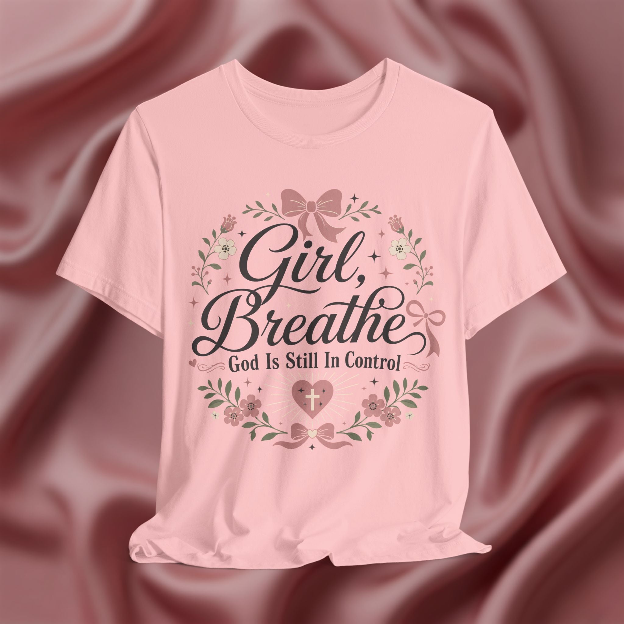 A light pink short-sleeve t-shirt with a graphic print reading 'Girl, Breathe God Is Still In Control' in black script, surrounded by a floral wreath with pink bows and green leaves, displayed against a draped satin fabric background.