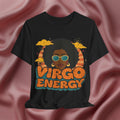 A black short-sleeve graphic T-shirt displayed against a draped dusty rose satin fabric background. The graphic features a retro-style illustration of a woman with an afro, sunglasses, and clouds, with the text 'VIRGO ENERGY' in orange and teal.