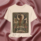 A soft cream crewneck t-shirt featuring a detailed vintage-style graphic of 'The Maiden Virgo' astrological card, displayed against a draped dusty rose satin background.